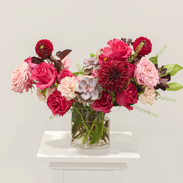 A dramatic and moody flower arrangement featuring deep red roses, burgundy dahlias, and succulents in a clear glass vase. This unique bouquet is perfect for those who appreciate rich colors and bold textures. Available for local delivery in Wake Forest, NC.
