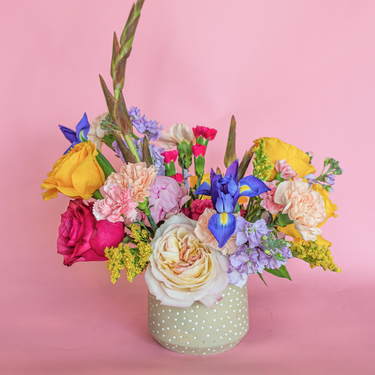 A vibrant and diverse flower arrangement featuring peach roses, yellow ranunculus, blue irises, pink carnations, and gladiolus. This eye-catching bouquet in a speckled vase is perfect for birthdays, anniversaries, or to send a cheerful greeting. Available for local delivery in Wake Forest, NC.