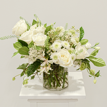 A serene arrangement of white and cream flowers, including roses, chrysanthemums, and delicate fillers, arranged in a clear glass vase. This elegant bouquet is perfect for sympathy occasions or to express condolences. Available for local delivery in Wake Forest, NC.