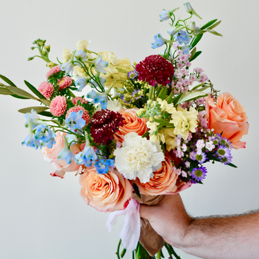 A colorful hand-tied bouquet with peach roses, blue delphinium, burgundy scabiosa, and a mix of wildflowers. This cheerful bouquet is perfect for birthdays, anniversaries, or just because. Available for local delivery in Wake Forest, NC.