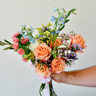 A hand-tied bouquet with peach roses, blue delphinium, and a mix of wildflowers and filler. This cheerful bouquet is perfect for birthdays, anniversaries, or to send a get-well wish. Available for local delivery in Wake Forest, NC.
