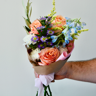 A hand-tied bouquet with peach roses, white carnations, and blue delphinium, accented with delicate fillers and greenery. This cheerful wrapped bouquet is perfect for birthdays, anniversaries, or to send a get-well wish. Available for local delivery in Wake Forest, NC.