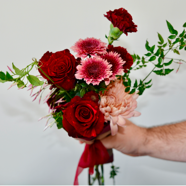 "A hand-tied bouquet featuring deep red roses, pink gerbera daisies, and carnations, accented with a peach chrysanthemum and greenery. This romantic bouquet is perfect for anniversaries or to express sympathy. Available for local delivery in Wake Forest, NC.